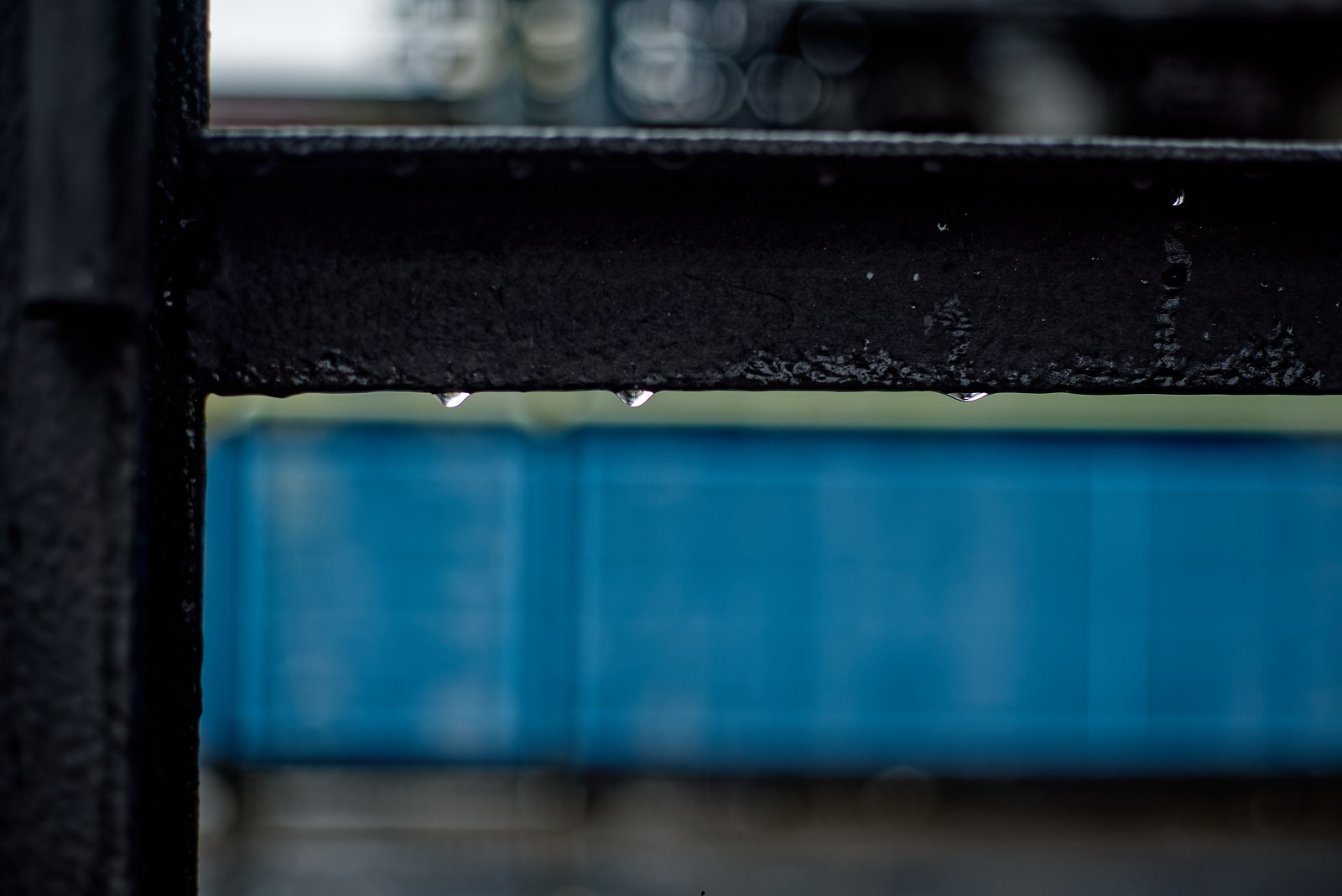 Metallener eckiger Handlauf mit einigen RegenTropfen, im hintergrund undefinierte blaue Fläche