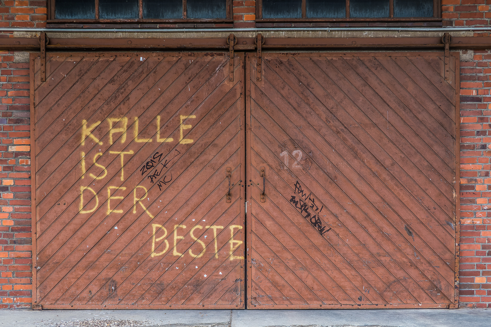 zweiflügeliges hölzernes Tor einer IndustrieHalle aus Backstein. Bekritzelt mit einigen unleserlichen Tags sowie groß und gelb "Kalle ist der Beste".