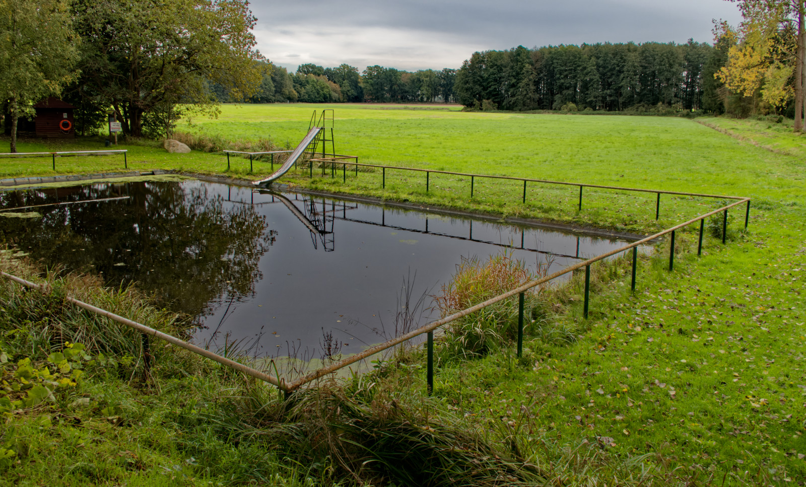 Ein Freibad oder rechteckig gefasster Teich mitten in einer Wiese,rundum umzäunt, auf einer Seite eine alte Rutsche ins Wasser.