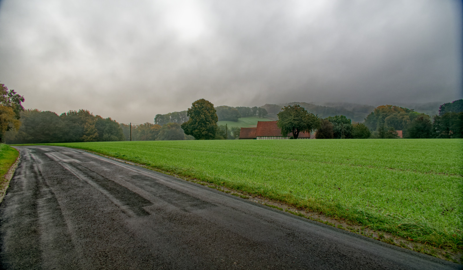 Eine feuchte, dunkle Landstraße unter dichten grauen Wolken, im Hintergrund grüne Hügel und ein rot gedecktes FachwerkHaus