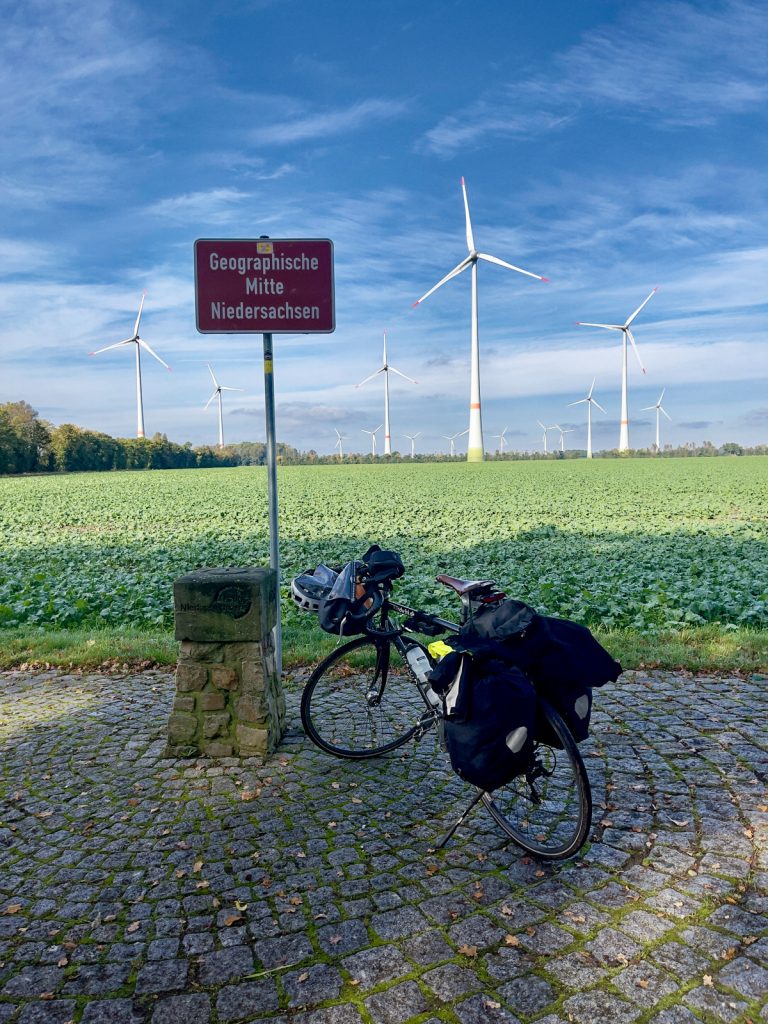 Ein gepflasterter Platz vor einem Feld und WindRädern. Auf dem Platz steht ein SteinMonument neben dem Schild "Geographische Mitte Niedersachsen", daneben noch ein Fahrrad.