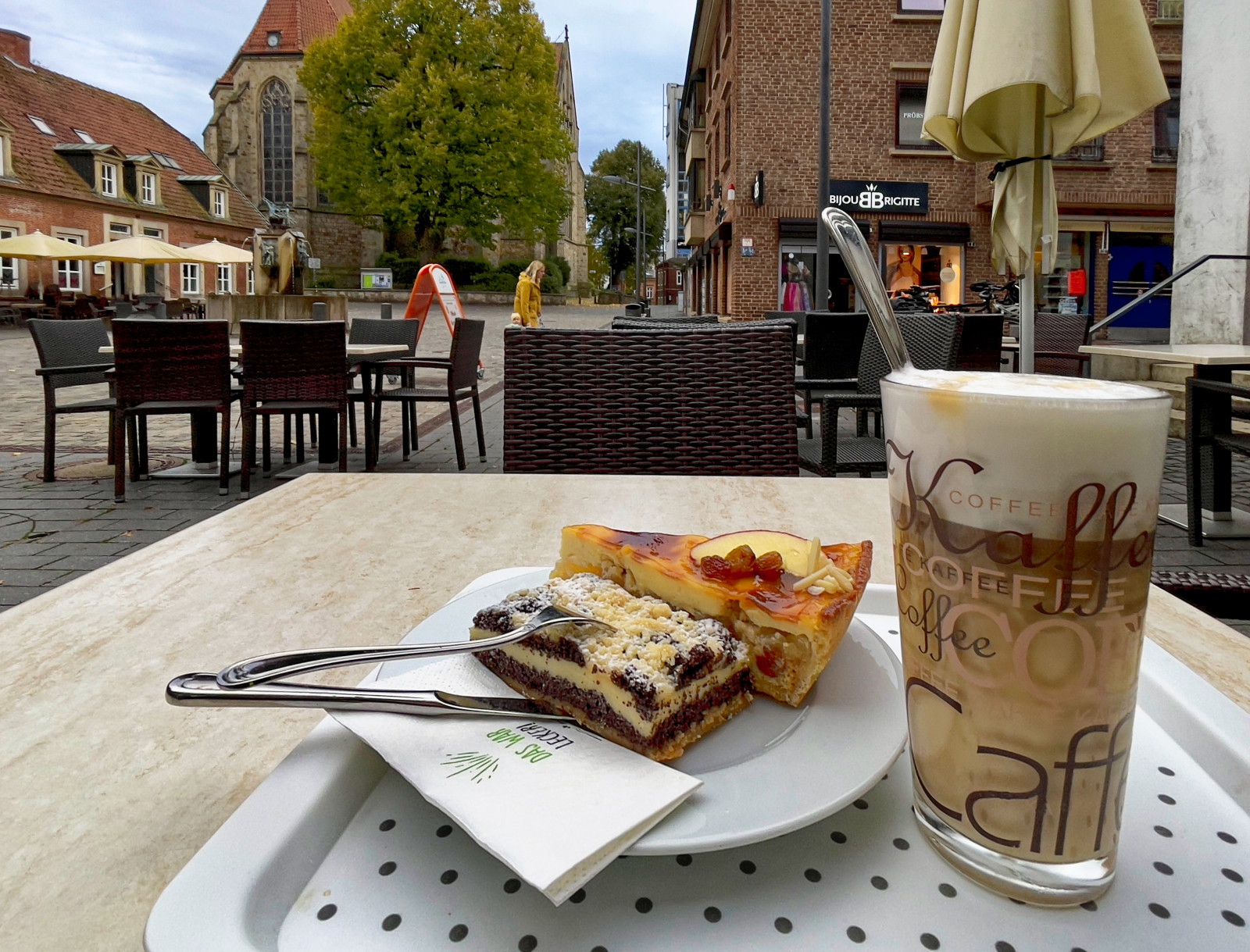 Ein Glas MichKaffee und zwei Stück Kuchen, Mohn und Apfel, auf einem Cafétisch auf einem weitläufigen städtischen Platz.