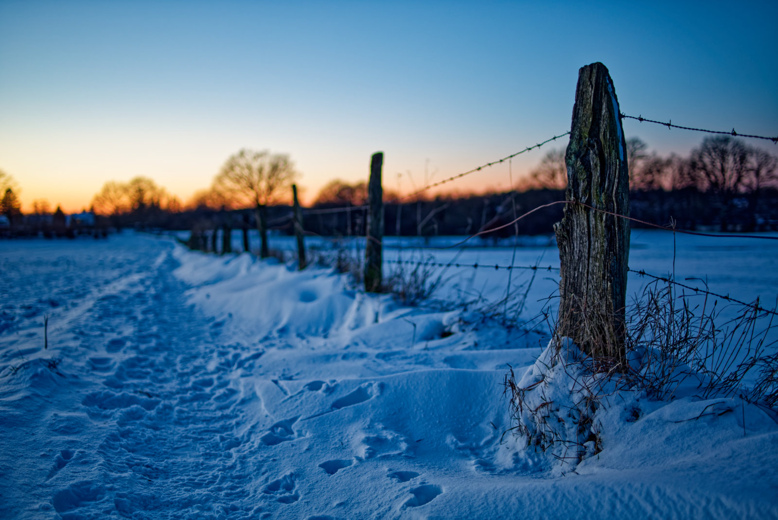 Blick längs eines DrahtZauns mit roh gehauenen Pfosten, an dem entlang sich Schnee angehäuft hat. Am Horizont rötliches Licht der untergehenden Sonne