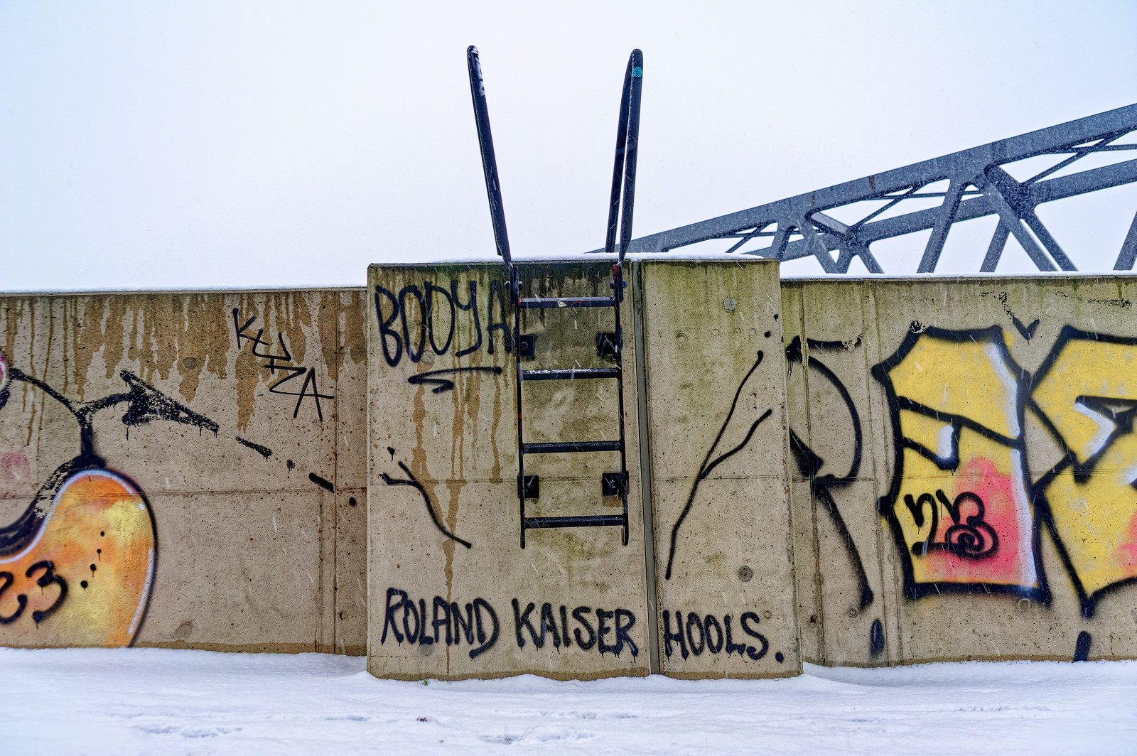 FlutSchutzWand mit einer kleinen schwarzen Leiter. Viele Tags von Graffiti Artists. Lesbar unter der Leiter: "Roland Kaiser Hools".