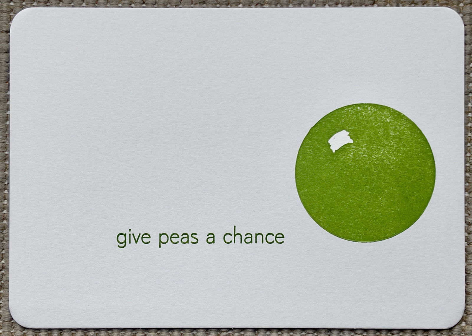 Minimalistische Postkarte: Auf natur-weißem Karton ist ein grüner Kreis gedruckt. Er hat eine kleine Fläche oben ausgespart, die einen räumlichen Eindruck vermittelt, als sei es der Licht-Reflex auf einer Kugel. Daneben, klein in magerer moderner Schrift, auch grün: „give peas a chance“ (Gib Erbsen eine Chance). Das ist ein Wort-Spiel mit dem Gleich-Klang aus peas und peace (Frieden).