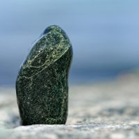 Ein dunkel-grüner, fein geaderter glatter Stein steht aufrecht wie eine Skulptur auf einem helleren Felsen vor dem unscharfen Hintergrund des blauen Himmels, es könnte auch das Meer sein.