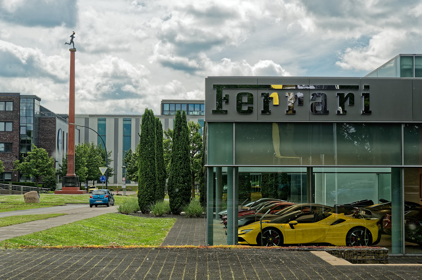 IndustrieGebiet unter wolkigem Himmel. Dominant der voll verglaste AusstellungsRaum eines Ferrari-Händlers, im VorderGrund eine gelbe, offensichtlich elektrische "Flunder", die an einem Kabel hängt.