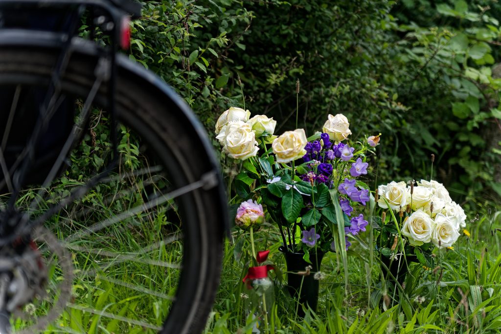 In hohem Gras stehen zwei schwarze BlumenVasen und eine gläserne mit roter Schleife. In zweien sind weiße Rosen und einige kleine lila Blümchen, in der gläsernen eine einzelne PfingstRose. Im VorderGrund sieht man unscharf ein einzelnes HinterRad eines FahrRades.