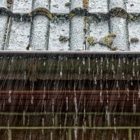 In engem Bildwinkel ist eine RegenRinne am Dach eines geklinkerte Hauses zu sehen. Es fällt starker Regen, und die Rinne ist bereits voll und es tropft über die Ränder.