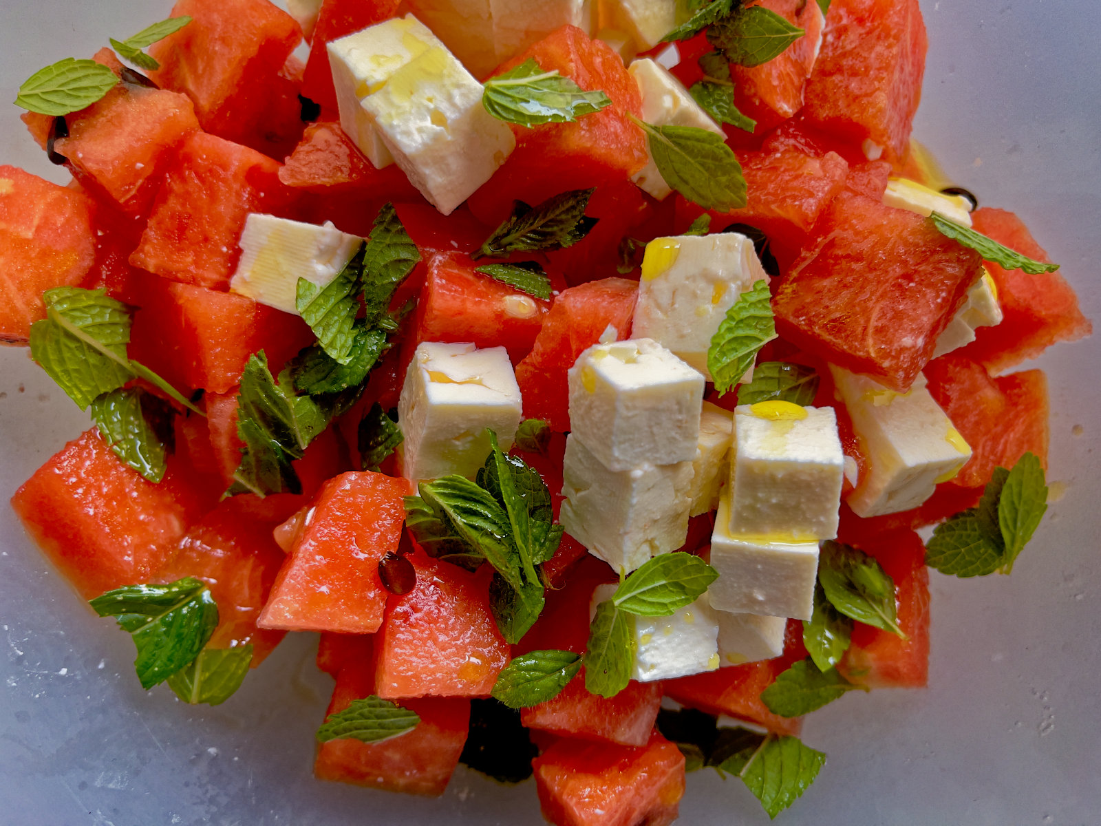 gewürfelte rote Melone und weißer Käse, gelbe Tröpfchen von Öl und kleine Blättchen verschiedener Kräuter in einer hellen Schale.