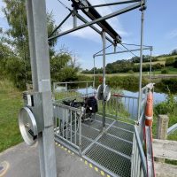 Ein GitterKorb, in dem ein Fahrrad mit Gepäck lehnt, ist wie eine SeilBahn-Gondel an einem Draht befestigt. Dieser Draht führt über einen Fluss. An einem Mast am SeilEnde ist eine Kurbel befestigt, auch in dem Korb selbst gibt es zwei Kurbeln.