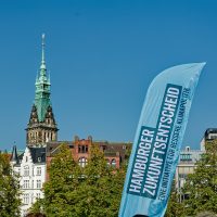 Quadratisches Foto: Vor klarem blauem Himmel der Turm des Hamburger Rathauses hinter verschiedenen Giebeln, rechts vorn eine Fahne mit dem Logo des Hamburger ZukunftsEntscheids.